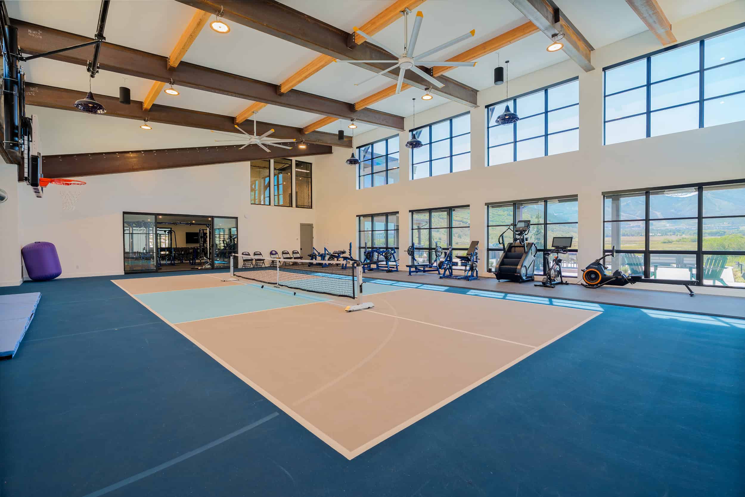 Bright indoor luxury home gym featuring a pickleball court, workout gear, and panoramic mountain views through large windows.